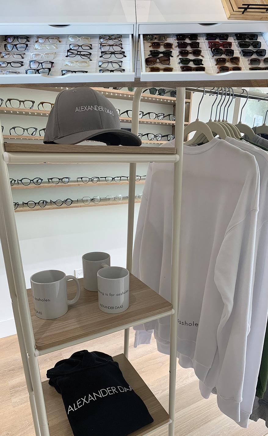 Alexander Daas frames line the shelves at Alexander Daas Eyewear & Opticians. At bottom, branded merchandise including the ‘Squinting is for assholes’ line.